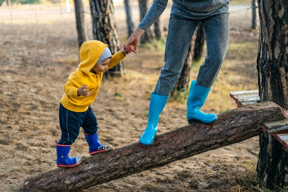 totschooling - mother holding toddler's hand as they walk up a fallen log in rain boots