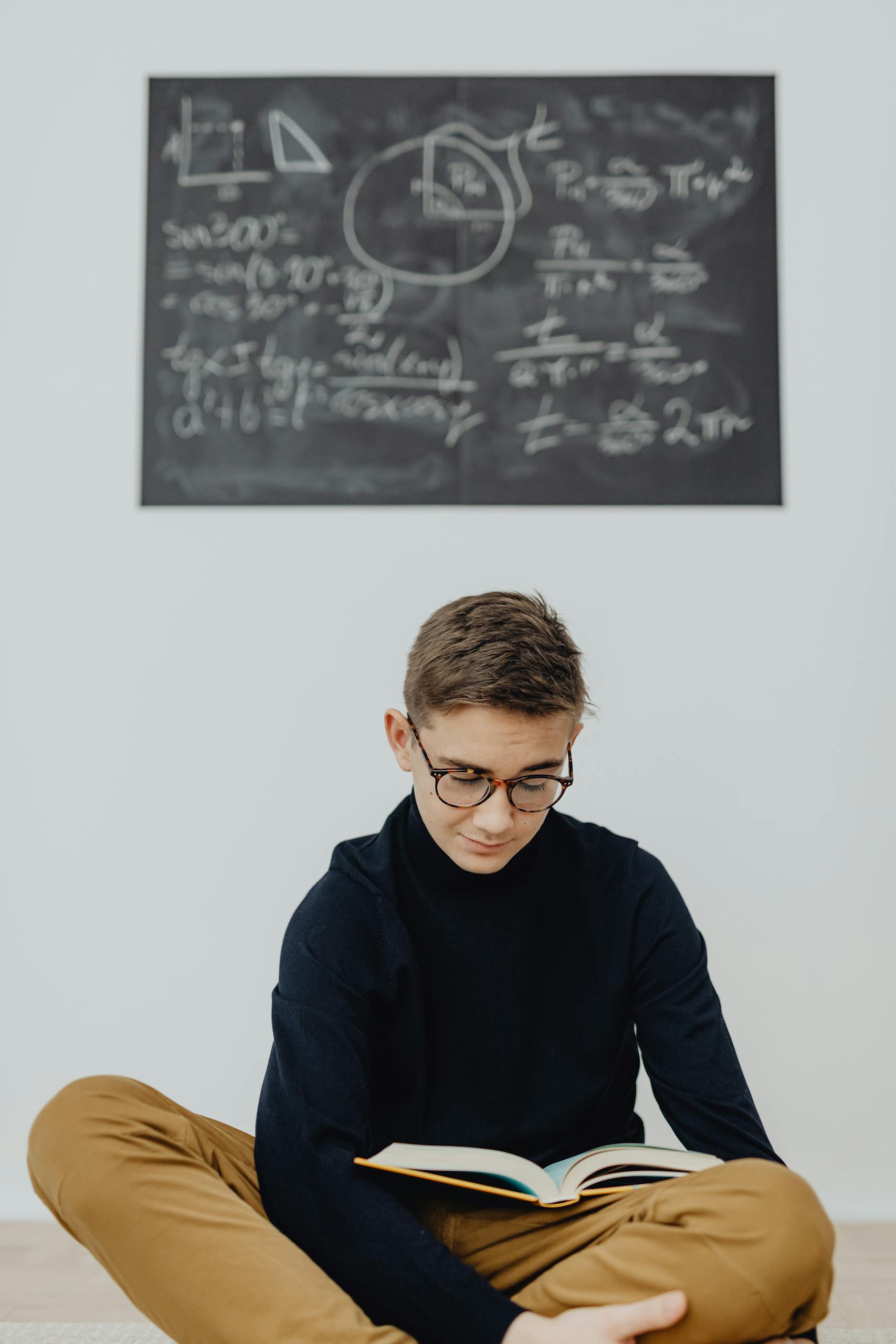 Teenage boy reading in front of chalkboard with math equations. Studio setting, educational theme.